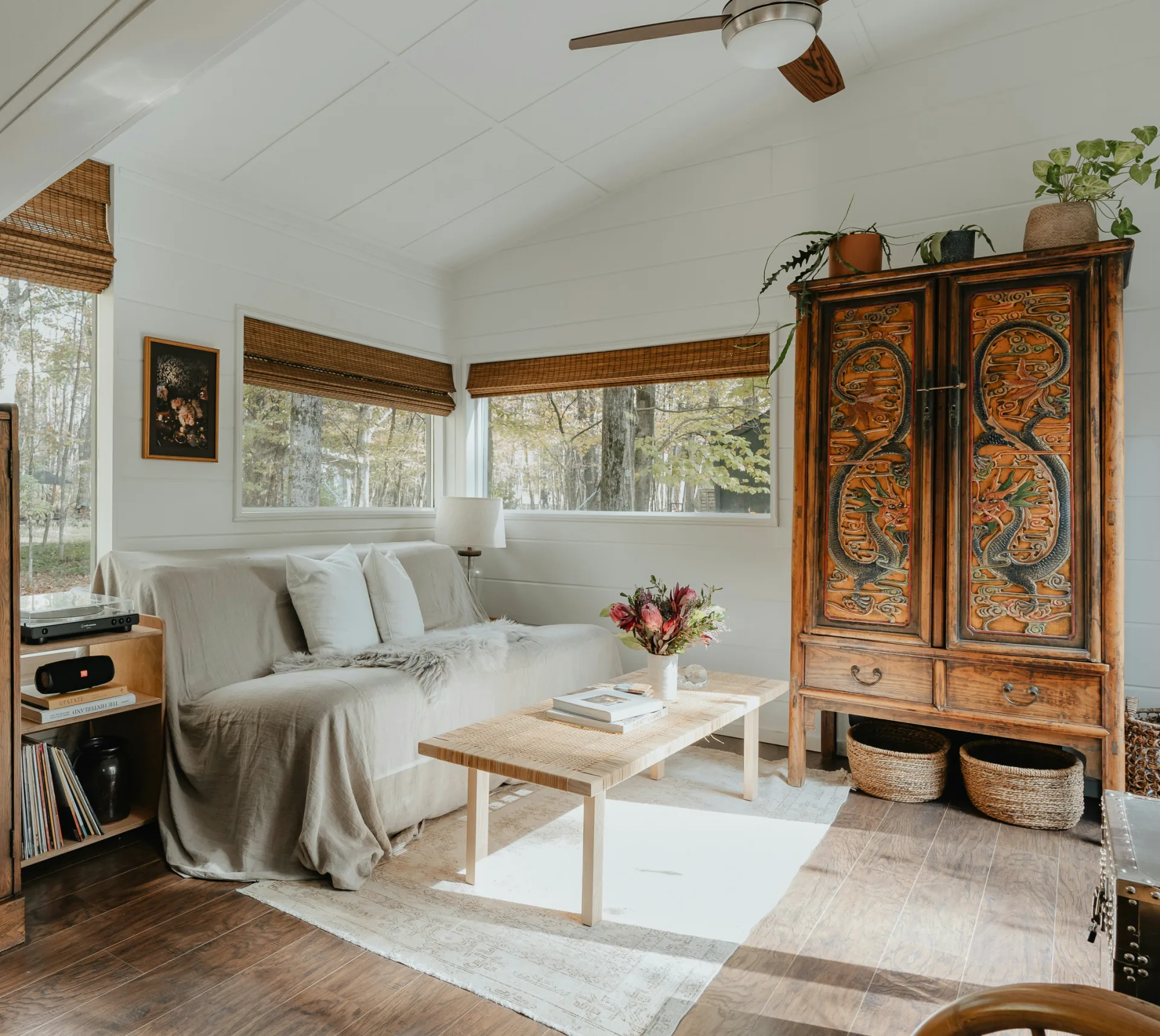 A bright living room with a carved wooden cabinet, a linen-covered sofa and bamboo blinds